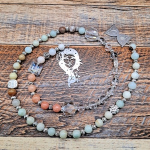 May include: A beaded prayer chain with a silver charm featuring a woman's face. The beads are a mix of light green, brown, and pink. The chain is on a wooden surface.