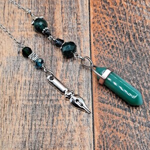 May include: Two silver-toned pen-themed pendants with dark green and black beads. One pendant features a pen nib design, the other a green crystal point with a rose gold cap. The pendants hang on delicate silver chains.