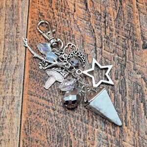 May include: Silver-toned keychain with various charms, including a star, moon, mushroom, and crystal beads. A light blue and white triangular pendant hangs from the bottom. The background is a wooden surface.