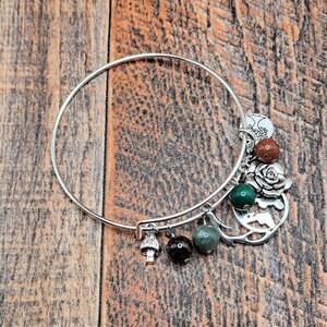 May include: A silver-colored bangle bracelet with various charms. Charms include a mushroom, dark and light green beads, a rose, a globe, and a white and brown stone. The bracelet is displayed on a wooden surface.