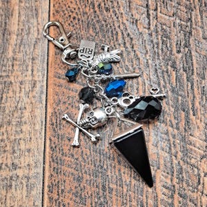 May include: A silver keychain with various charms, including a skull, crossbones, a black triangular pendant, and blue and black beads. The keychain also features a small RIP charm and a heart-shaped charm.