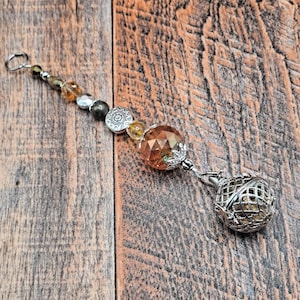 May include: A decorative charm with a silver-colored cage pendant and a faceted amber-colored crystal bead. The charm also features a series of smaller beads in shades of gold, silver, and black.