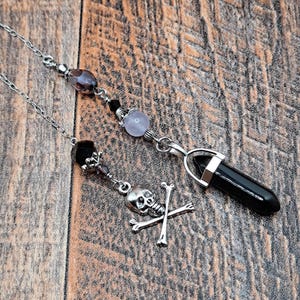 May include: Two silver pendulums with chains. One features a skull and crossbones charm, the other a black crystal point. Both have beaded accents in shades of purple, black, and clear.