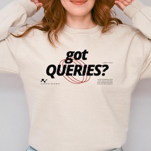 May include: A beige long-sleeve sweatshirt featuring the black text "got QUERIES?" centered on the chest. The design includes red circular line graphics, small black checkered icons, and additional small black text details below the main graphic.