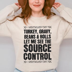 May include: A beige crewneck sweatshirt featuring black distressed text that reads: TURKEY, GRAVY, BEANS & ROLLS LET ME SEE THE SOURCE CONTROL, framed by diagonal striped lines at the top and bottom of the graphic design on the front of the garment.