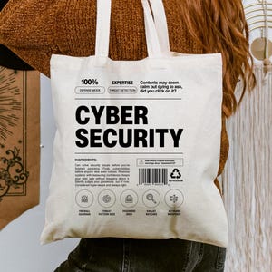 May include: A cream-coloured tote bag with the words "CYBER SECURITY" in bold black letters. The bag also features additional text and icons related to cybersecurity. The bag is being held by a person.