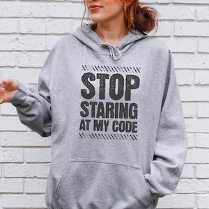 Könnte beinhalten: Ein graumelierter Kapuzenpullover mit einer Kängurutasche und einer Kapuze mit Kordelzug. Auf der Brust befindet sich der schwarze Schriftzug "STOP STARING AT MY CODE" in einer fetten, abgenutzten Schrift, eingerahmt von zwei diagonalen schwarzen Strichen.