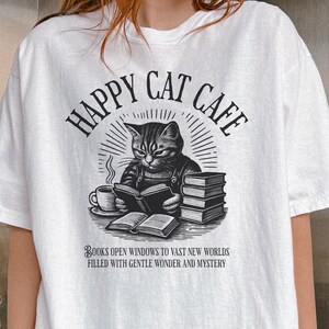 Model wearing oversized white colored Comfort Colors® T-Shirt with a graphic of a vintage cat reading a book with a cup of coffee and book stack nearby, graphic is styled with Happy Cat Cafe headline and a quirky lost in translation-inspired subhead.