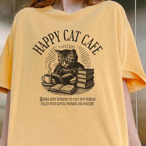 Model wearing oversized mustard colored Comfort Colors® T-Shirt with a graphic of a vintage cat reading a book with a cup of coffee and book stack nearby, graphic is styled with Happy Cat Cafe headline and a quirky lost in translation-inspired subhead.
