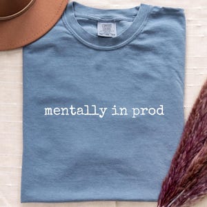 May include: A blue jean colored t-shirt with the text mentally in prod printed in white across the chest. The shirt features a crew neckline and a Comfort Colors brand label at the inner collar. A brown hat and dried pampas grass are visible.