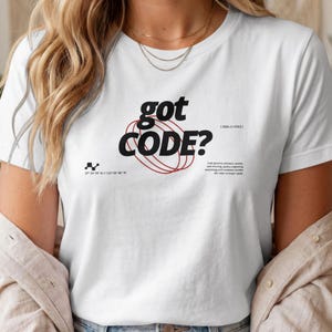 Op de afbeelding: Een wit t-shirt met korte mouwen en de zwarte tekst "got CODE?" in het midden op de borst. Het ontwerp bevat een rode cirkelvormige afbeelding, kleine zwarte coördinatentekst en extra kleine zwarte tekstblokken onder de afbeelding.