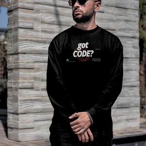 Model wearing a black colored Gildan G18000 crewneck sweatshirt with a retro-tech graphic design featuring the phrase GOT CODE? in bold typography over red orbital linework with small technical details and humorous token-themed microtext.
