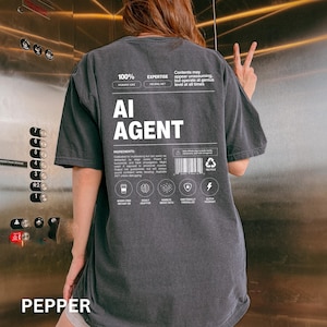May include: A dark gray t-shirt with the text "AI AGENT" in white. The shirt has additional text and graphics, including "100% HUMAN-LIKE" and "EXPERTISE NEURAL NET". The shirt is a casual, oversized fit.