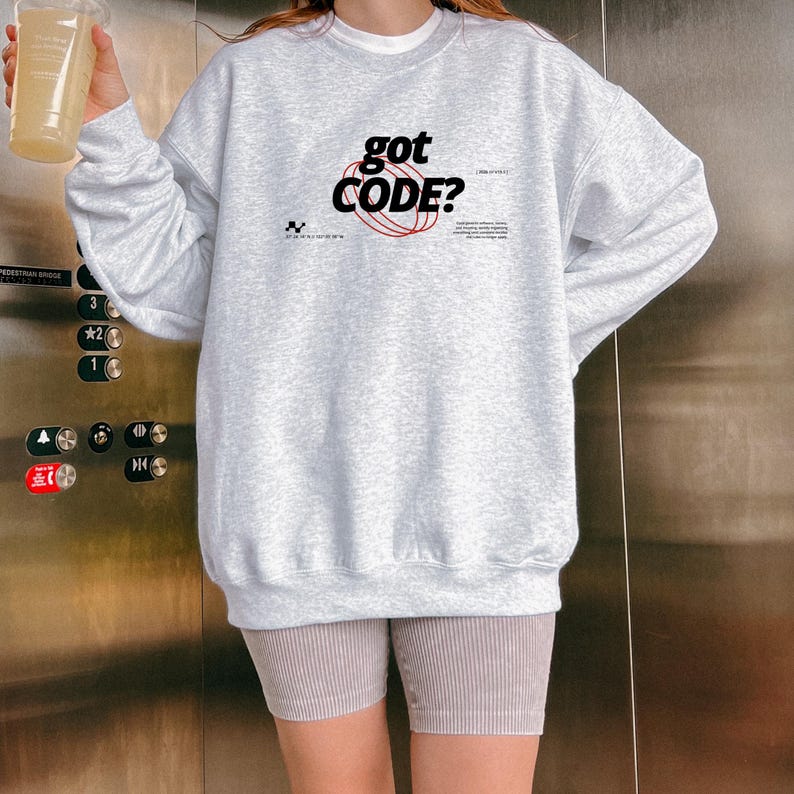 Model wearing an ash colored Gildan G18000 crewneck sweatshirt with a retro-tech graphic design featuring the phrase GOT CODE? in bold typography over red orbital linework with small technical details and humorous token-themed microtext.