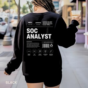 May include: Black sweatshirt with the text "SOC ANALYST" in white. The sweatshirt has additional text and graphics, including "100% EAGLE-EYED" and "EXPERTISE SEM SENSED". The bottom of the sweatshirt says "BLACK".