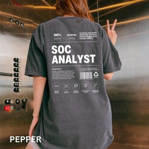 May include: A dark gray t-shirt with the text "SOC ANALYST" in large white letters. The shirt features additional white text and graphics, including "100% EAGLE-EYED" and "EXPERTISE SEM SENSE". The word "PEPPER" is at the bottom.