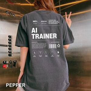 Model is wearing a pepper colored t-shirt shown from the back, displaying an AI Trainer parody label graphic designed like a tech product spec sheet with alignment indicators, ingredients, and dry humor.
