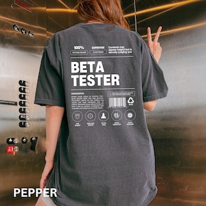 May include: A dark gray t-shirt with the text "BETA TESTER" in large white letters. The shirt also features smaller text and graphics, including "100% PATTERN AWARE" and "EXPERTISE FLAW FINDER". The word "PEPPER" is at the bottom.
