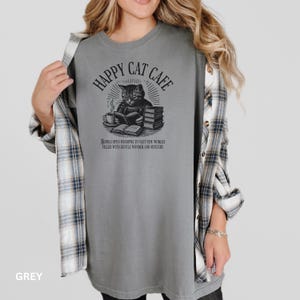Model wearing oversized grey colored Comfort Colors® T-Shirt with a graphic of a vintage cat reading a book with a cup of coffee and book stack nearby, graphic is styled with Happy Cat Cafe headline and a quirky lost in translation-inspired subhead.