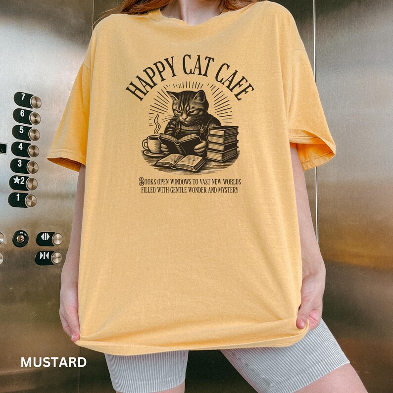 Model wearing oversized mustard colored Comfort Colors® T-Shirt with a graphic of a vintage cat reading a book with a cup of coffee and book stack nearby, graphic is styled with Happy Cat Cafe headline and a quirky lost in translation-inspired subhead.