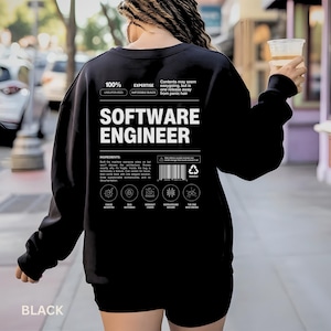 May include: A black long-sleeve sweatshirt featuring white text on the back that reads SOFTWARE ENGINEER, along with a nutrition-style label design including the words 100% EXPERTISE, INGREDIENTS, and various small icons with labels like CHAOS INVENTOR.