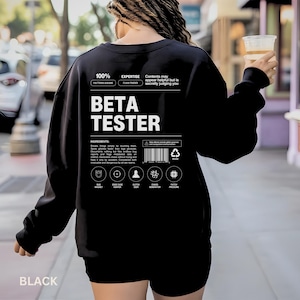 May include: Black sweatshirt with the text "BETA TESTER" in white. The sweatshirt has additional text and graphics, including "100% PATTERN AWARE" and "EXPERTISE FLAW FINDER". The word "BLACK" is at the bottom.