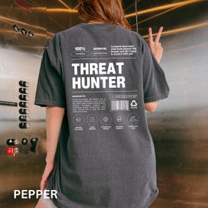 May include: A charcoal gray t-shirt featuring white graphic text on the back. The design includes the words THREAT HUNTER, a barcode, and various icons with labels like ARTIFACT RECOVERER, CURSE BREAKER, ATTACK PATH TRACKER, MALWARE EXORCIST, and RABBIT-HOLE GUIDE.