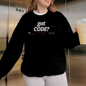Model wearing a black colored Gildan G18000 crewneck sweatshirt with a retro-tech graphic design featuring the phrase GOT CODE? in bold typography over red orbital linework with small technical details and humorous token-themed microtext.