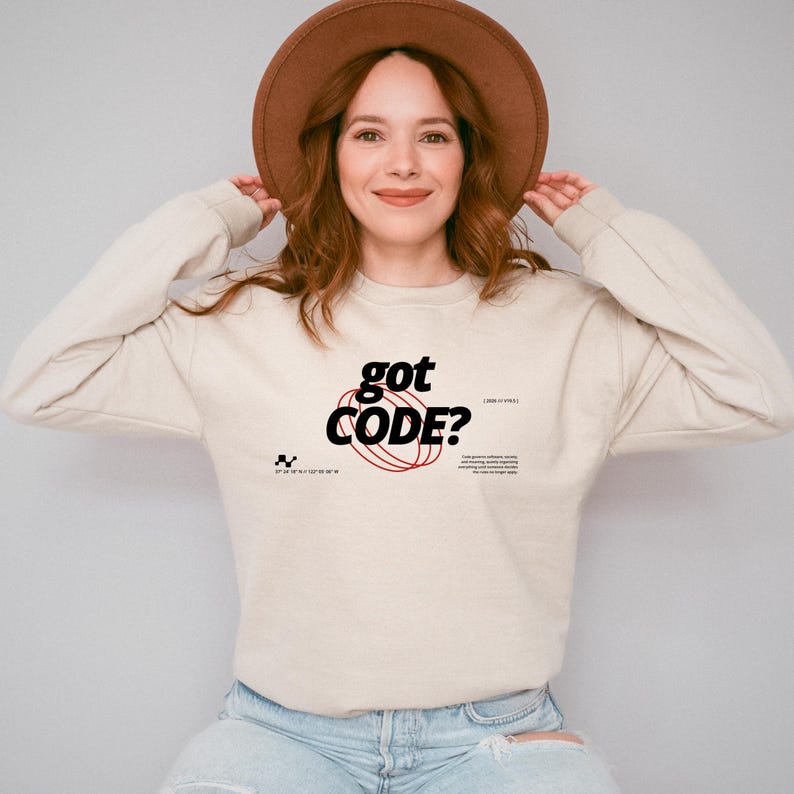 Model wearing a sand colored Gildan G18000 crewneck sweatshirt with a retro-tech graphic design featuring the phrase GOT CODE? in bold typography over red orbital linework with small technical details and humorous token-themed microtext.