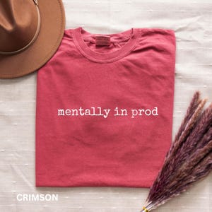 Może przedstawiać: Karmazynowy t-shirt z kr&oacute;tkim rękawem z białym napisem "mentally in prod" nadrukowanym na klatce piersiowej. Koszulka ma okrągły dekolt i jest pokazana obok brązowego kapelusza z szerokim rondem oraz wiązki suszonej fioletowej trawy.