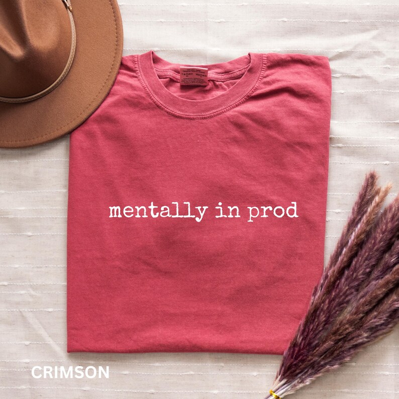 Może przedstawiać: Karmazynowy t-shirt z kr&oacute;tkim rękawem z białym napisem "mentally in prod" nadrukowanym na klatce piersiowej. Koszulka ma okrągły dekolt i jest pokazana obok brązowego kapelusza z szerokim rondem oraz wiązki suszonej fioletowej trawy.