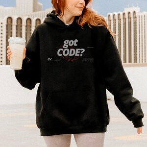 Model wearing a black colored Gildan G18500 pullover hoodie with a retro-tech graphic design featuring the phrase GOT CODE? in bold typography over red orbital linework with small technical details and humorous token-themed microtext.
