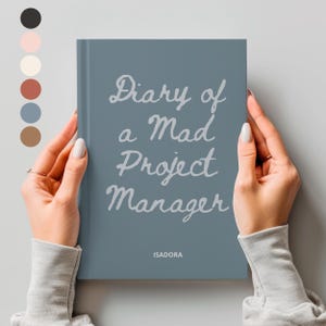 May include: A slate blue hardcover journal with the text Diary of a Mad Project Manager in a light gray script font on the front cover. The name ISADORA is printed in a small, simple sans-serif font at the bottom center of the book cover.