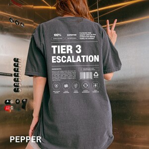 May include: Dark gray t-shirt with the text "TIER 3 ESCALATION" in large white letters. The shirt also features smaller text and graphics, including "100% OVERCLOCKED" and "EXPERTISE THE IMPOSSIBLE". The word "PEPPER" is at the bottom.