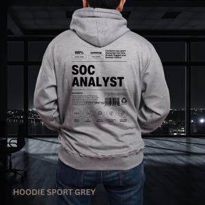 May include: A gray hoodie with the text "SOC ANALYST" printed on the back. Additional text includes "100% Eagle-Eyed" and "Expertise: Sixth Sense". The hoodie is labeled "HOODIE SPORT GREY".