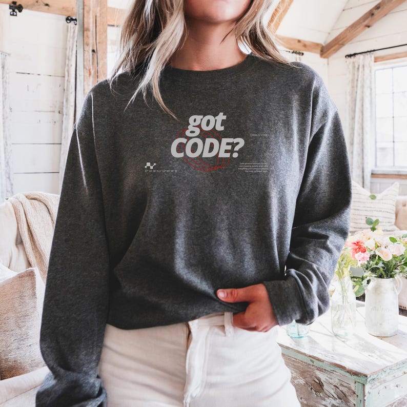 Model wearing a dark heather colored Gildan G18000 crewneck sweatshirt with a retro-tech graphic design featuring the phrase GOT CODE? in bold typography over red orbital linework with small technical details and humorous token-themed microtext.
