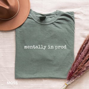 Może przedstawiać: Szałwiowo-zielony t-shirt z okrągłym dekoltem i białym napisem "mentally in prod" wykonanym czcionką przypominającą maszynę do pisania. Koszulka leży obok brązowego kapelusza z szerokim rondem oraz wiązki suszonej trawy pampasowej w odcieniu r&oacute;żu.