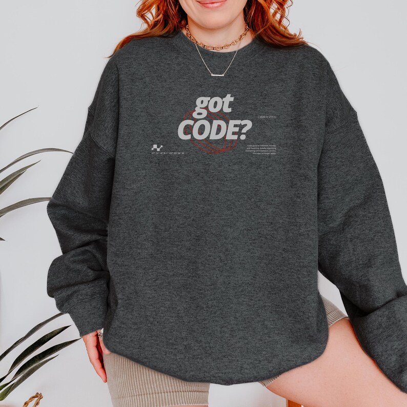 Model wearing a dark heather colored Gildan G18000 crewneck sweatshirt with a retro-tech graphic design featuring the phrase GOT CODE? in bold typography over red orbital linework with small technical details and humorous token-themed microtext.