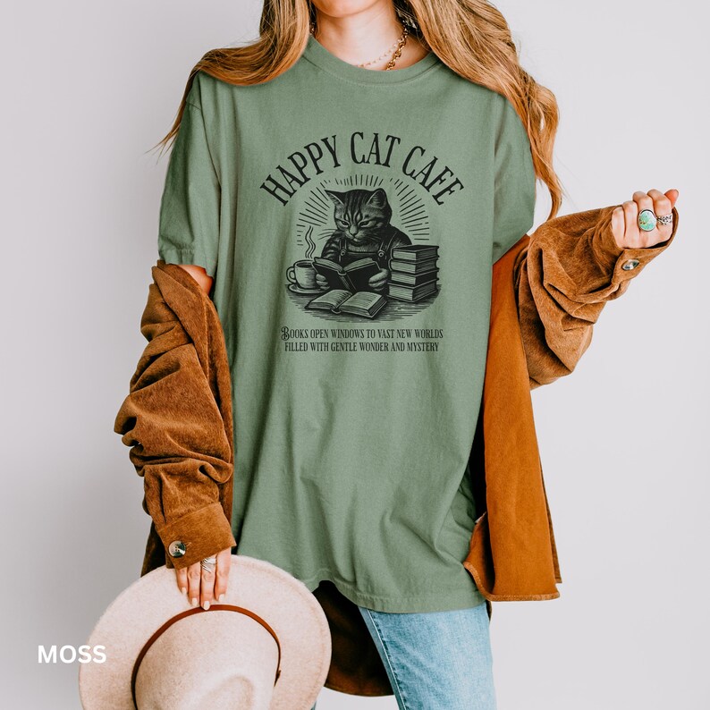 Model wearing oversized moss colored Comfort Colors® T-Shirt with a graphic of a vintage cat reading a book with a cup of coffee and book stack nearby, graphic is styled with Happy Cat Cafe headline and a quirky lost in translation-inspired subhead.