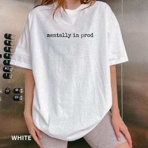 Może przedstawiać: Biały t-shirt z kr&oacute;tkim rękawem z czarnym napisem "mentally in prod" nadrukowanym na klatce piersiowej. Odzież posiada okrągły dekolt oraz luźny, swobodny kr&oacute;j, wykonana z gładkiej białej tkaniny o prostym i przejrzystym wzornictwie.