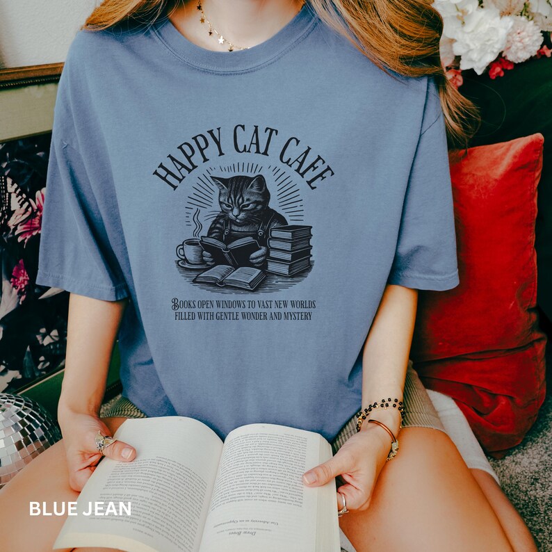 Model wearing oversized blue jean colored Comfort Colors® T-Shirt with a graphic of a vintage cat reading a book with a cup of coffee and book stack nearby, graphic is styled with Happy Cat Cafe headline and a quirky lost in translation-inspired subhead.