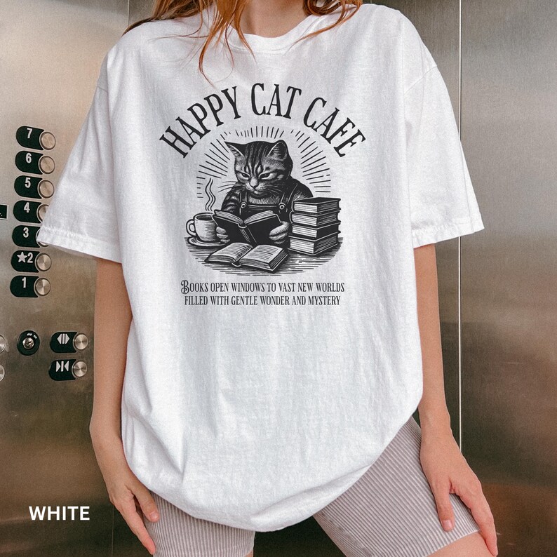 Model wearing oversized white colored Comfort Colors® T-Shirt with a graphic of a vintage cat reading a book with a cup of coffee and book stack nearby, graphic is styled with Happy Cat Cafe headline and a quirky lost in translation-inspired subhead.