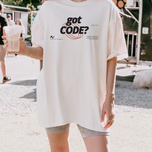 Model wearing an ivory Comfort Colors 1717 T-shirt with a retro-tech GOT CODE? graphic featuring bold black typography, red orbital linework, and small technical details.