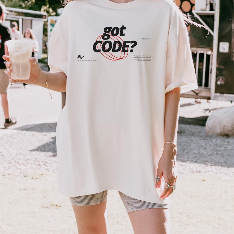 Model wearing an ivory Comfort Colors 1717 T-shirt with a retro-tech GOT CODE? graphic featuring bold black typography, red orbital linework, and small technical details.