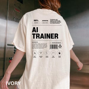 Model is wearing an ivory t-shirt shown from the back, displaying an AI Trainer parody label graphic designed like a tech product spec sheet with alignment indicators, ingredients, and dry humor.