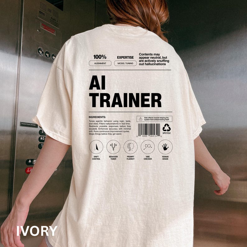 Model is wearing an ivory t-shirt shown from the back, displaying an AI Trainer parody label graphic designed like a tech product spec sheet with alignment indicators, ingredients, and dry humor.