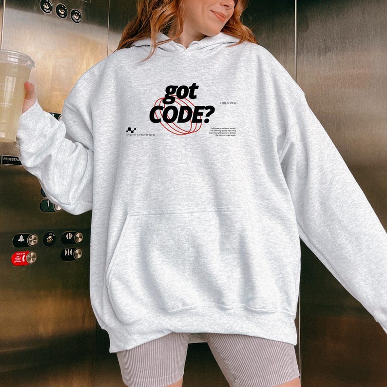 Model wearing an ash colored Gildan G18500 pullover hoodie with a retro-tech graphic design featuring the phrase GOT CODE? in bold typography over red orbital linework with small technical details and humorous token-themed microtext.