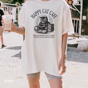 Model wearing oversized ivory colored Comfort Colors® T-Shirt with a graphic of a vintage cat reading a book with a cup of coffee and book stack nearby, graphic is styled with Happy Cat Cafe headline and a quirky lost in translation-inspired subhead.