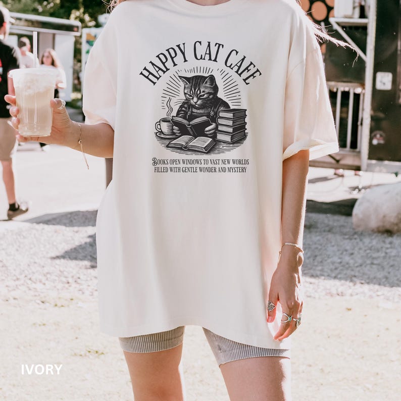 Model wearing oversized ivory colored Comfort Colors® T-Shirt with a graphic of a vintage cat reading a book with a cup of coffee and book stack nearby, graphic is styled with Happy Cat Cafe headline and a quirky lost in translation-inspired subhead.
