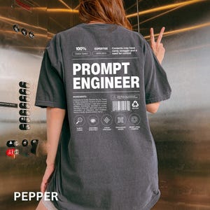 May include: Dark gray t-shirt with the text "PROMPT ENGINEER" in large white letters on the back. The shirt also features smaller text and graphics, including the word "INGREDIENTS" and a barcode. The shirt is a casual, oversized fit.